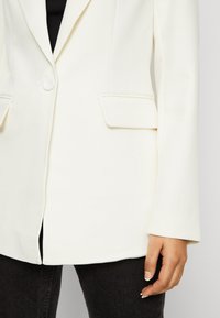 Bílý blazer s klopou, jedním knoflíkem a dvěma předními kapsami. Hladká textura, přiléhavý střih a dlouhé rukávy.