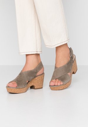 Pies que llevan sandalias de cuña de ante taupe con plataformas de corcho y dedos al aire, combinadas con pantalones blancos cortos.
