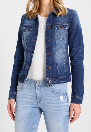 Frau trägt eine blaue Jeansjacke mit Knöpfen über einem weißen Spitzen-Top und hellblauen zerrissenen Jeans, steht mit entspannten Händen.