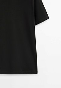 T-shirt noir à manches courtes présenté sur fond blanc, montrant une vue latérale et partielle de l'avant du vêtement.