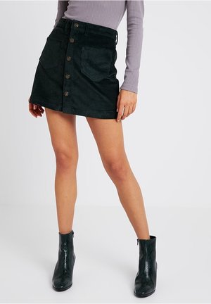 Femme portant une jupe noire en velours côtelé à boutons sur le devant, un haut violet clair à manches longues côtelé et des bottines noires texturées.