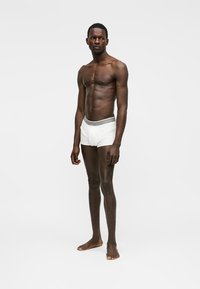 Homme mince debout, pieds nus, portant un boxer blanc avec une ceinture à motifs, sur fond uni.