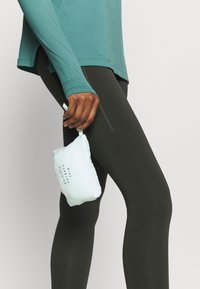 Blusa de manga larga color azul verde y leggings oscuros; una mano sostiene una mochila de color azul claro con texto negro. Cuenta con un bolsillo con cremallera en la pierna.