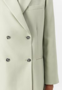 Dobbeltspent blazer i lysegrønn stoff, med mørke knapper, en frontlomme og en strukturert silhuett med klaffekrage.