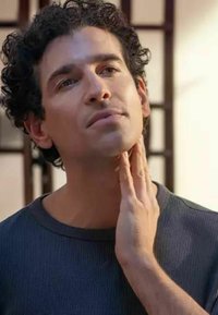 Uomo con capelli ricci che indossa una maglietta blu a coste, toccandosi la mascella, con illuminazione soffusa e uno sfondo sfocato.
