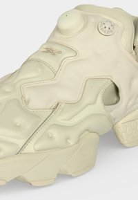 Chaussure de sport de couleur crème avec semelle texturée, fermetures à boutons ronds et détails de design angulaires sur la partie supérieure.