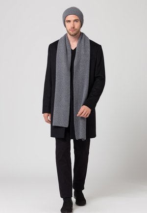 Beanie in maglia grigia e sciarpa a coste abbinata, indossati con un cappotto nero e pantaloni neri. Design semplice, texture morbida e colori neutri.