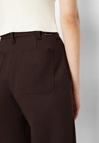 Gros plan d'une personne portant un pantalon marron foncé avec poche arrière et un haut sans manches côtelé crème, debout devant un fond clair uni.