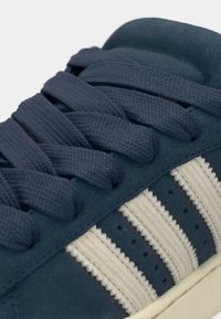 Ναυτικό σουέτ αθλητικό παπούτσι με ανοιχτό μπεζ ριγέ λεπτομέρειες, υφασμένα κορδόνια και διατρητή διακόσμηση στα πλάγια. Στρογγυλερός σχεδιασμός μύτης.