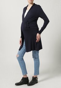 Námořnický pletený kardigan s uvazováním, dlouhými rukávy a kapucí. Nositý přes bílý top, kombinovaný se světle modrými roztrhanými džínami a černými kotníkovými botami.