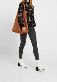 Braune Wildleder-Umhängetasche, schwarze Bluse mit floralem Muster, graue Skinny Jeans, weiße Ankle Boots mit quadratischen Zehen und Holzabsätzen.