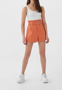 Shorts ajustés orange avec une ceinture dotée d'une boucle circulaire, en tissu lisse, associés à un débardeur blanc côtelé et des baskets blanches.