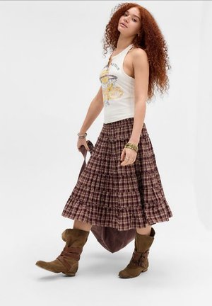 Jonge vrouw met krullend rood haar die een wit tanktopje, een geruite midirok en bruine laarzen draagt, loopt met een tas in de hand tegen een effen achtergrond.
