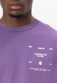 Fialové bavlněné tričko s bílým potiskem textu: "OBEY," "AROUND THE GLOBE," a názvy měst. Kulatý výstřih a krátké rukávy.