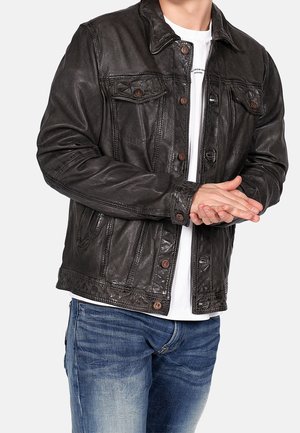 Hombre con chaqueta de cuero negra sobre camisa blanca y vaqueros azules, con las manos entrelazadas delante.