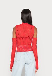 Red sleeveless top with a high neck; features a textured pattern of cut-outs and fitted long sleeves. Paired with light blue jeans.