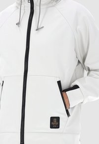 Giacca bianca con chiusura a zip frontale, cordini grigi e dettagli neri. Presenta un'etichetta con logo e tasche laterali. Tessuto liscio e resistente all'acqua.