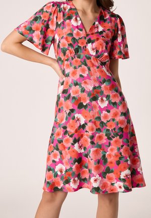 Donna che indossa un vestito rosa al ginocchio con stampa floreale arancione e bianca e maniche corte svolazzanti, posando con una mano sui fianchi.