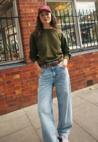 Jongere in olijfgroene trui, lichtblauwe wijde jeans, rode pet en sneakers, staand met handen in de zakken bij een bakstenen muur en ramen.