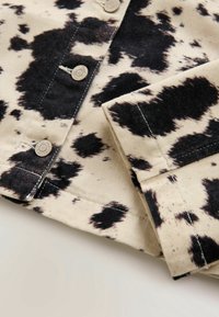 Tissu beige clair avec motif irrégulier à l'imprimé vache noire, boutons-pression métalliques et coutures visibles sur un revers de manche plié.