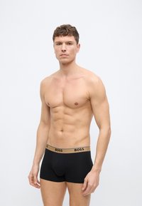 Άνδρας χωρίς πουκάμισο στέκεται μπροστά σε απλό φόντο, φοράει μαύρα μποξέρ με χρυσή μέση με την επιγραφή «BOSS», κοιτάζοντας προς τα εμπρός με ουδέτερη έκφραση.