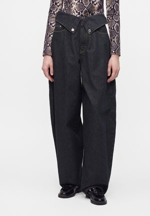 Vrouw draagt zwarte high-waist jeans met wijde pijpen en een bijzondere overlappende gulp met knopen, gecombineerd met zwarte loafers en een top met slangenprint.