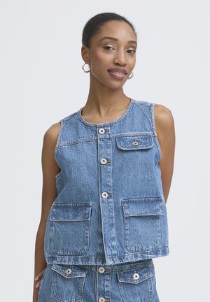 Kobieta z zaczesanymi do tyłu włosami, nosząca srebrne kolczyki-koła, bezrękawnik z niebieskiego denimu z kieszeniami i guzikami oraz pasującą spódnicę z denimu.