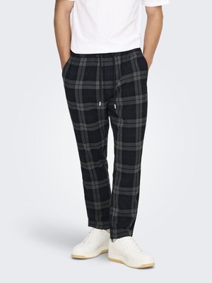 Bărbat purtând pantaloni negri și gri în carouri cu șnur, adidași albi și o cămașă albă cu mânecă scurtă, stând cu mâinile în buzunare.