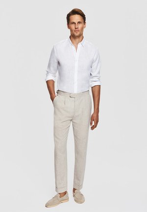 Mężczyzna stoi z rękami w kieszeniach, ubrany w białą koszulę zapinaną na guziki, beżowe spodnie i beżowe mokasyny na jednolitym tle.