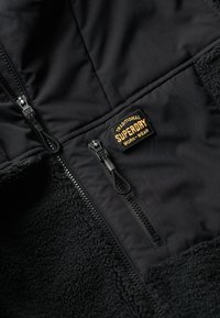 Sort fleecejakke med en robust lynlås, blød tekstur og et kontraststofpanel. Inkluderer et "Superdry Work-Wear" mærke.
