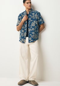 Camicia a maniche corte blu con motivi floreali bianchi, abbinata a pantaloni a gamba larga color crema e scarpe slip-on grigie. Il modello è in piedi dritto.