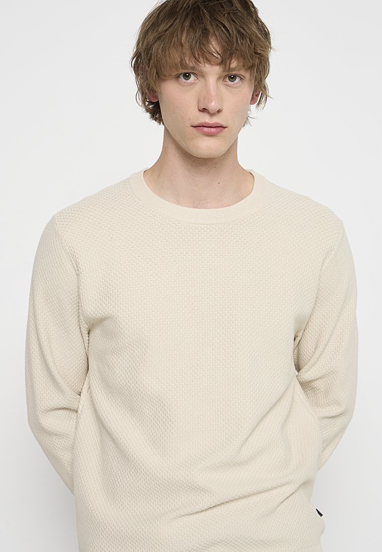 Ljusbeige stickad tröja med ett texturerat mönster, rund halsringning och långa ärmar. Har en svart logotyp-tag i nederkant.