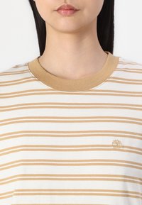 Striped t-shirt in white and beige, featuring a crew neck with a tan ribbed collar and a small, embroidered logo on the chest.