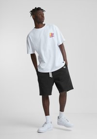 Hvit bomulls T-skjorte med et flerfarget grafisk logo på brystet, kombinert med svarte shorts og hvite joggesko.