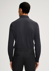 Camicia nera a maniche lunghe con una texture liscia, con polsini abbottonati e colletto classico, abbinata a pantaloni neri su misura con doppie tasche posteriori.