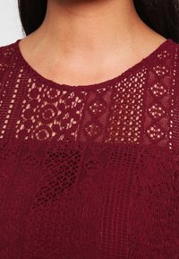 Burgundy topp med intrikata spetsdetaljer med olika mönster, inklusive geometriska former och blommotiv, samt en bred halsringning.