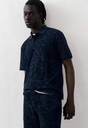 Ung mand med mørk hud og dreadlocks iført en marineblå mønstret poloshirt og matchende bukser mod en ensfarvet lys baggrund.