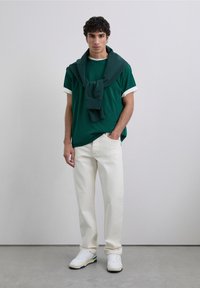 Junger Mann steht drinnen und trägt ein dunkelgrünes T-Shirt, weiße Jeans, weiße Sneakers und einen dunkelgrauen Sweatshirt, der über die Schultern gelegt ist.