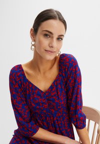 Abito con stampa floreale presenta uno sfondo blu con fiori rossi, una scollatura a V e maniche a tre quarti, completato da orecchini a cerchio dorati.