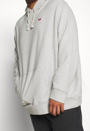 Person in einem hellgrauen Hoodie mit Kordeln und einem kleinen roten Levi's-Logo auf der Brust, kombiniert mit dunklen Hosen.