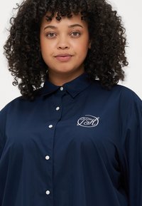 Marineblå skjorte med knappelukking og spiss krage, med en hvit brodert logo og fem hvite knapper foran.