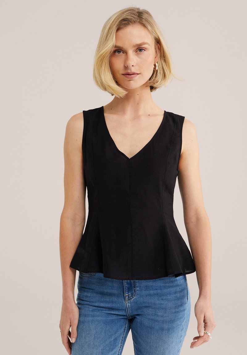 Femme blonde portant des boucles d'oreilles créoles dorées, un haut noir sans manches à col en V et à basque, et un jean bleu, sur un fond uni beige.