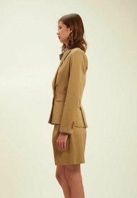 Costume ajusté couleur marron clair avec une veste cintrée et une jupe jusqu'aux genoux. Tissu texturé avec un design structuré et de subtils accents de boutons.