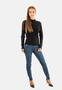 Pull en tricot noir à côtes avec un col haut, associé à un jean bleu slim et des escarpins noirs brillants. La mannequin se tient devant un fond blanc.
