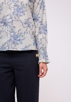 Blusa floreale grigia chiara con motivi blu e verdi, chiusura a bottoni e maniche arricciate. Abbinata a pantaloni blu scuro con dettagli dorati.