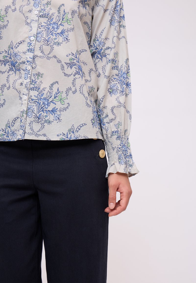 Blusa floreale grigia chiara con motivi blu e verdi, chiusura a bottoni e maniche arricciate. Abbinata a pantaloni blu scuro con dettagli dorati.