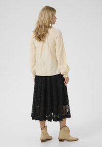 Blouse créme avec un motif texturé et des manches bouffantes, associée à une jupe en dentelle noire et des bottines beige. Accent sur le tissu et la silhouette.