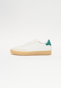 White sneaker with beige rubber sole, teal accent on heel, smooth leather upper, and flat white laces. Minimalistic design.