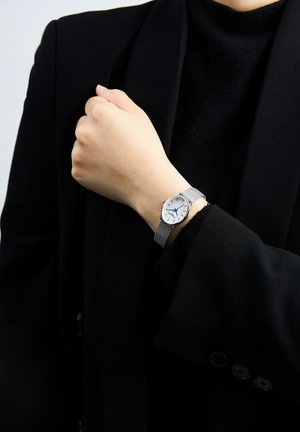 Montre-bracelet argentée avec un bracelet en maille, cadran blanc, aiguille des heures bleue et index noirs, portée sur une main contre un blazer noir.