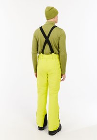 Skibroek in felgeel met zwarte bretels, gecombineerd met een langärmige olijfgroene shirt en een bijpassende beanie. Voorzien van ritszakken.